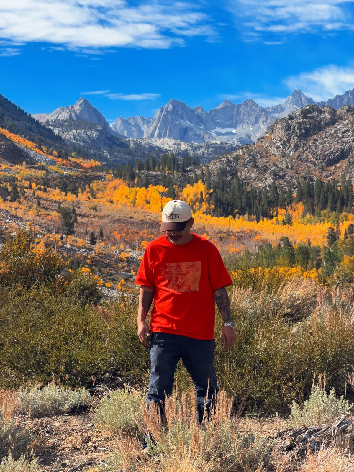 Standing in sagebrush in a mountain canyon, yellow aspens and granite peaks behind, wearing an orange tee and a cream cap, looking down.
